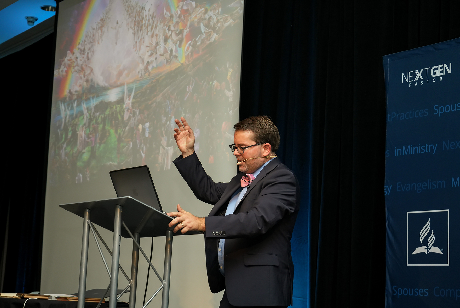 White man speaking with arm uplifted in front of a graphic slide depicting Jesus and the angels descending from heaven at the second coming. 