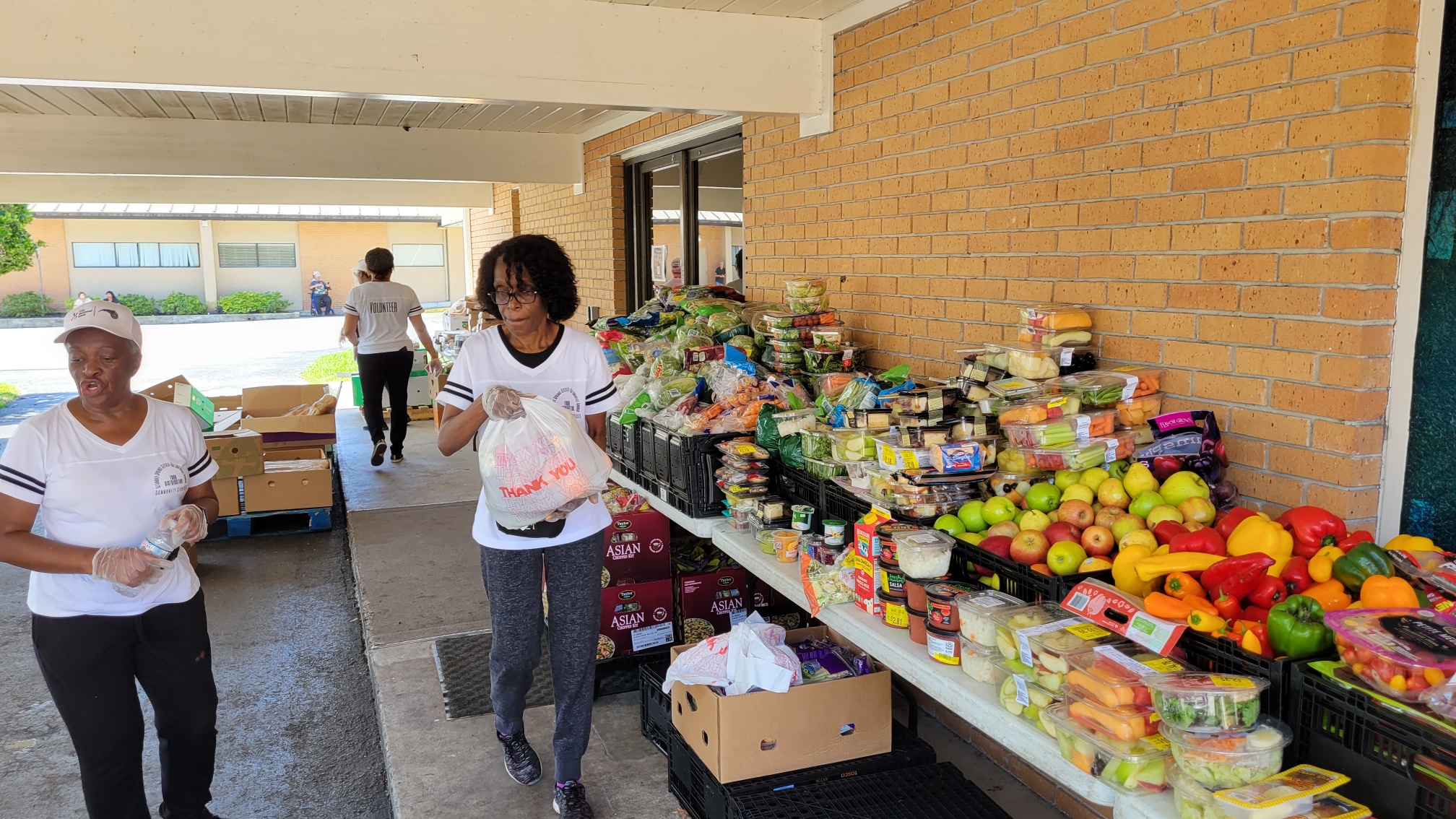 Ladies are standing in front of tables laden with fruits and vegetables at a food drive.