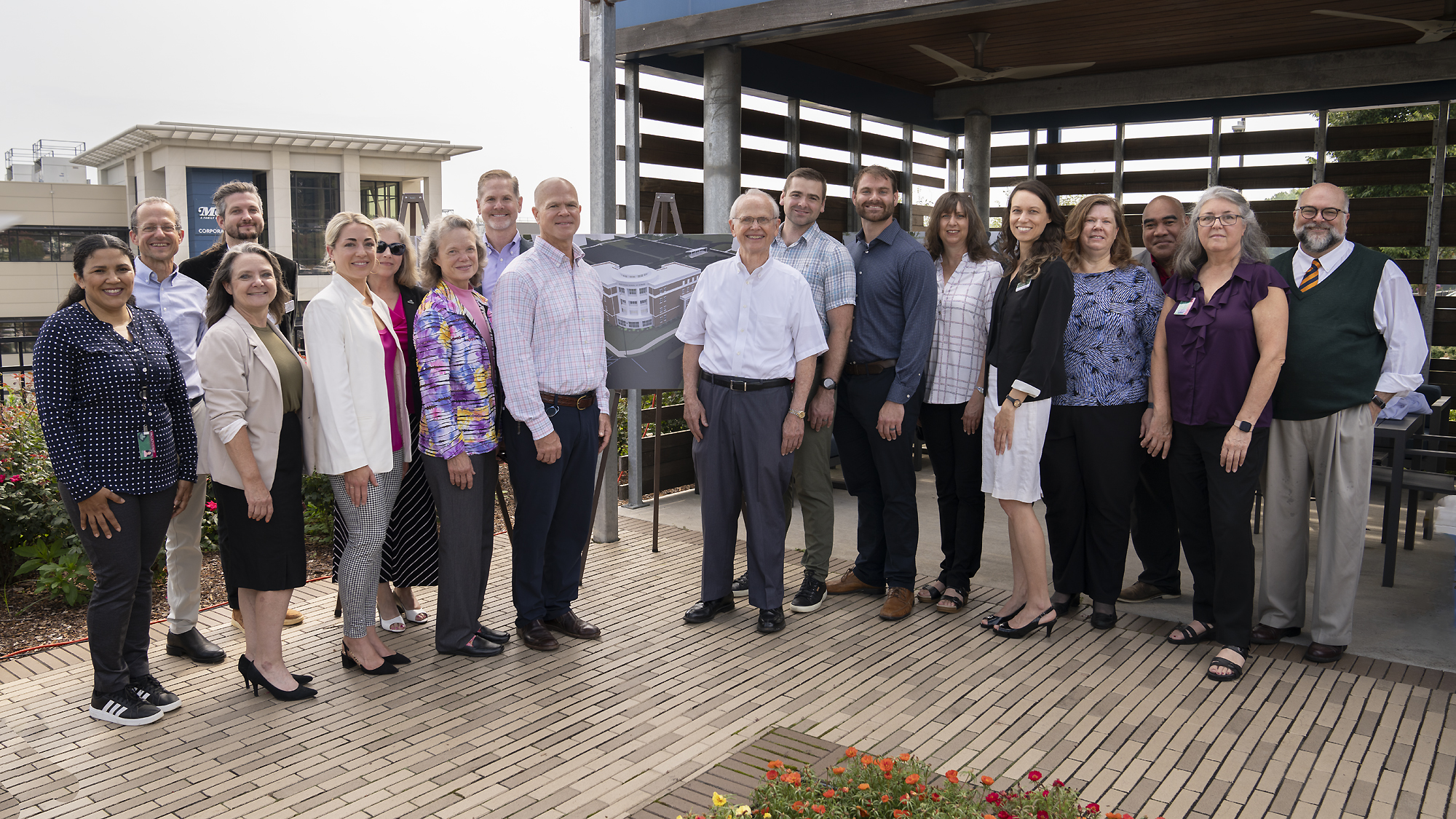 A group of smiling individuals stand in front of an outside structure with a rendering of a building in the middle of them