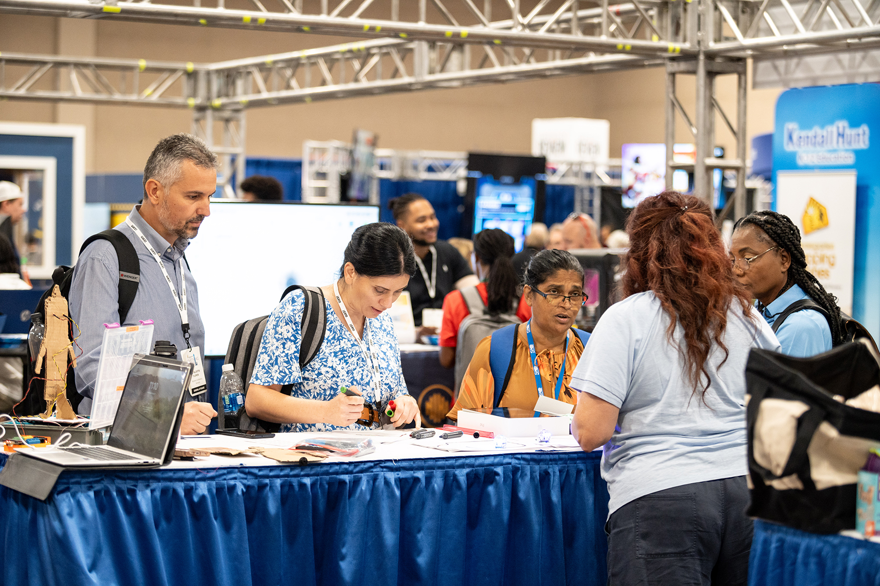 A woman behind a table speaks to three women and a man in a busy exhibit hall