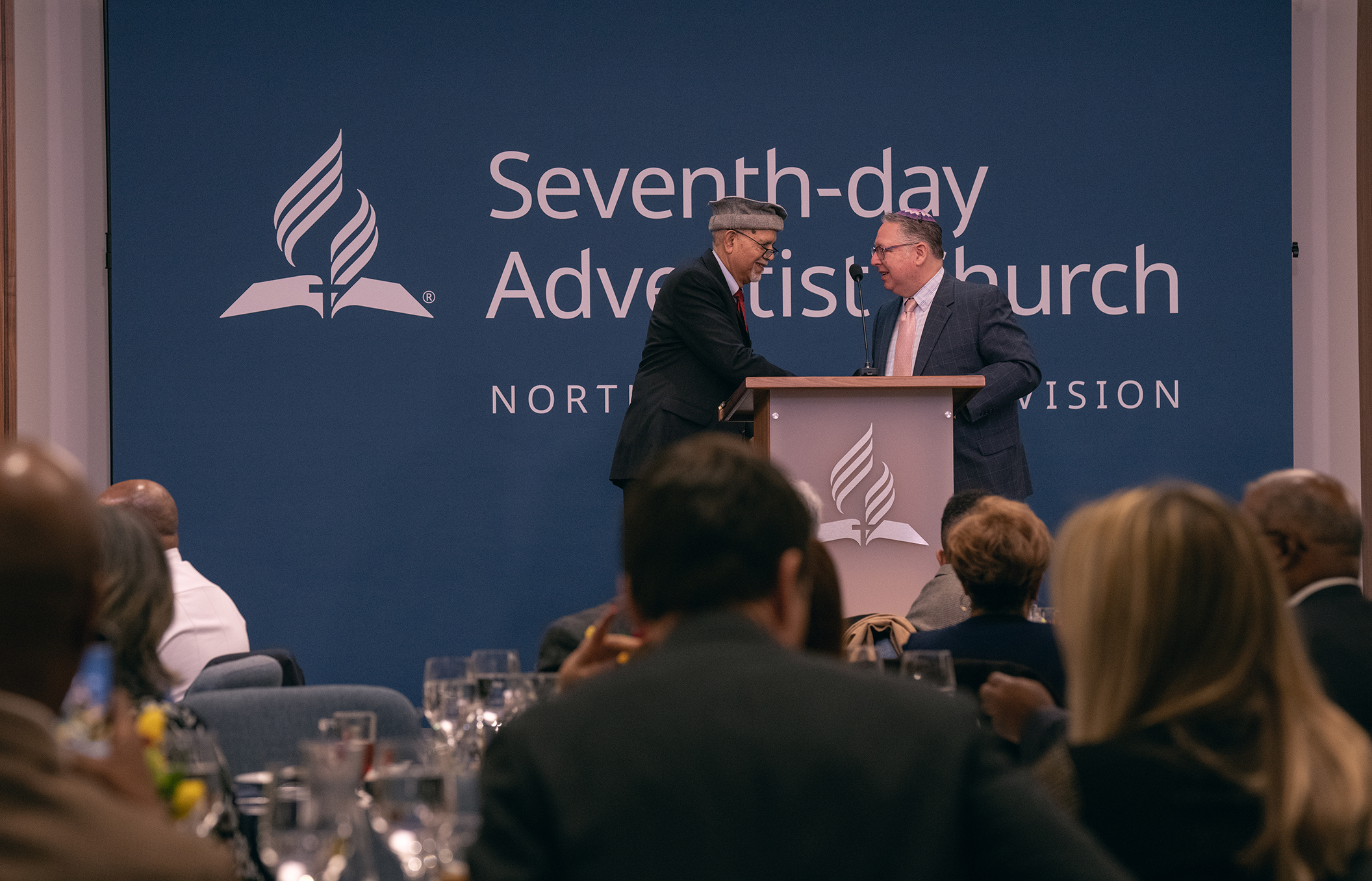 Rabbi and imam in religious costume shake hands behind a podium in a conference room. 