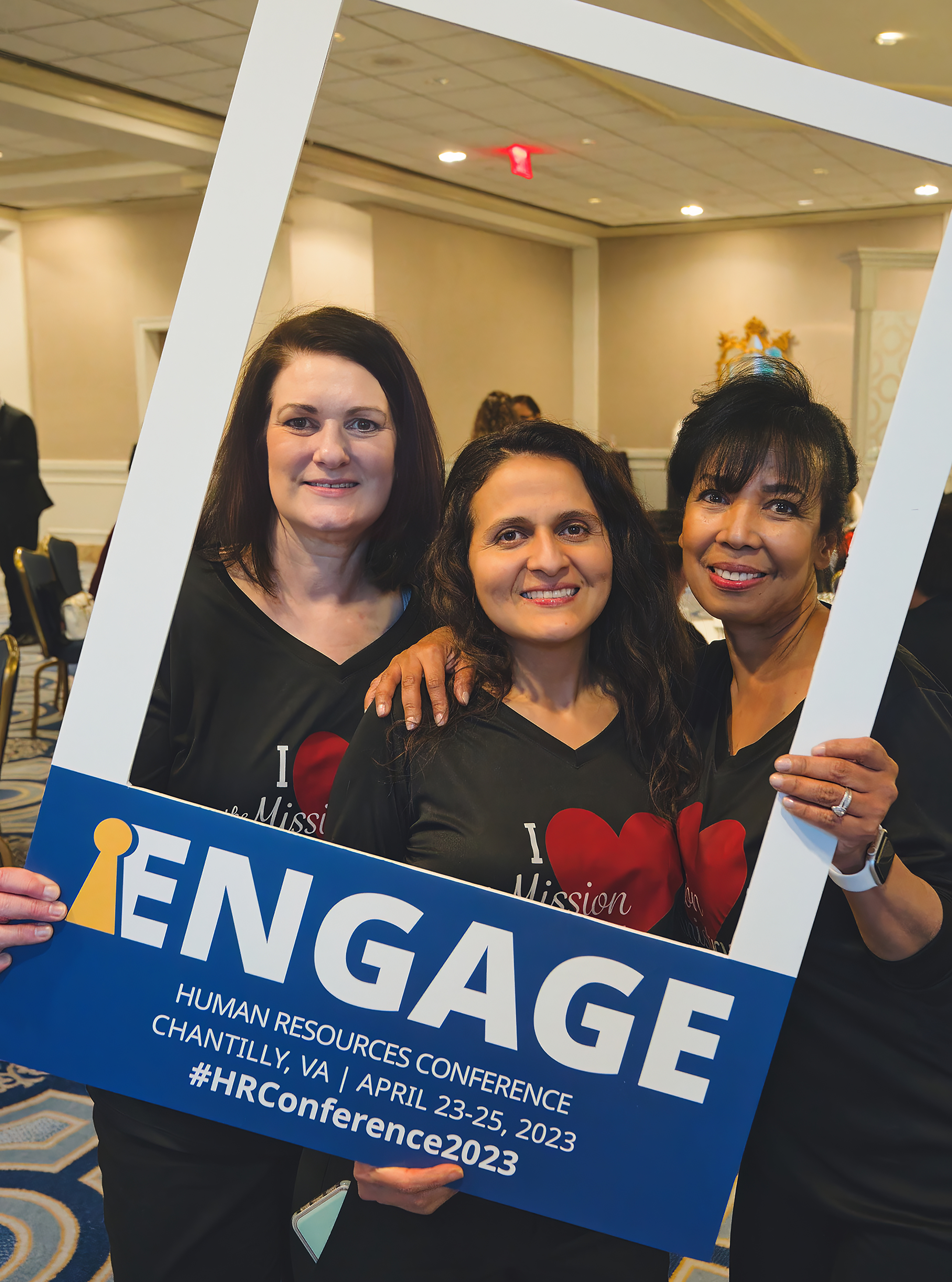 Three woman standing with a big cardboard frame that reads "Engage"