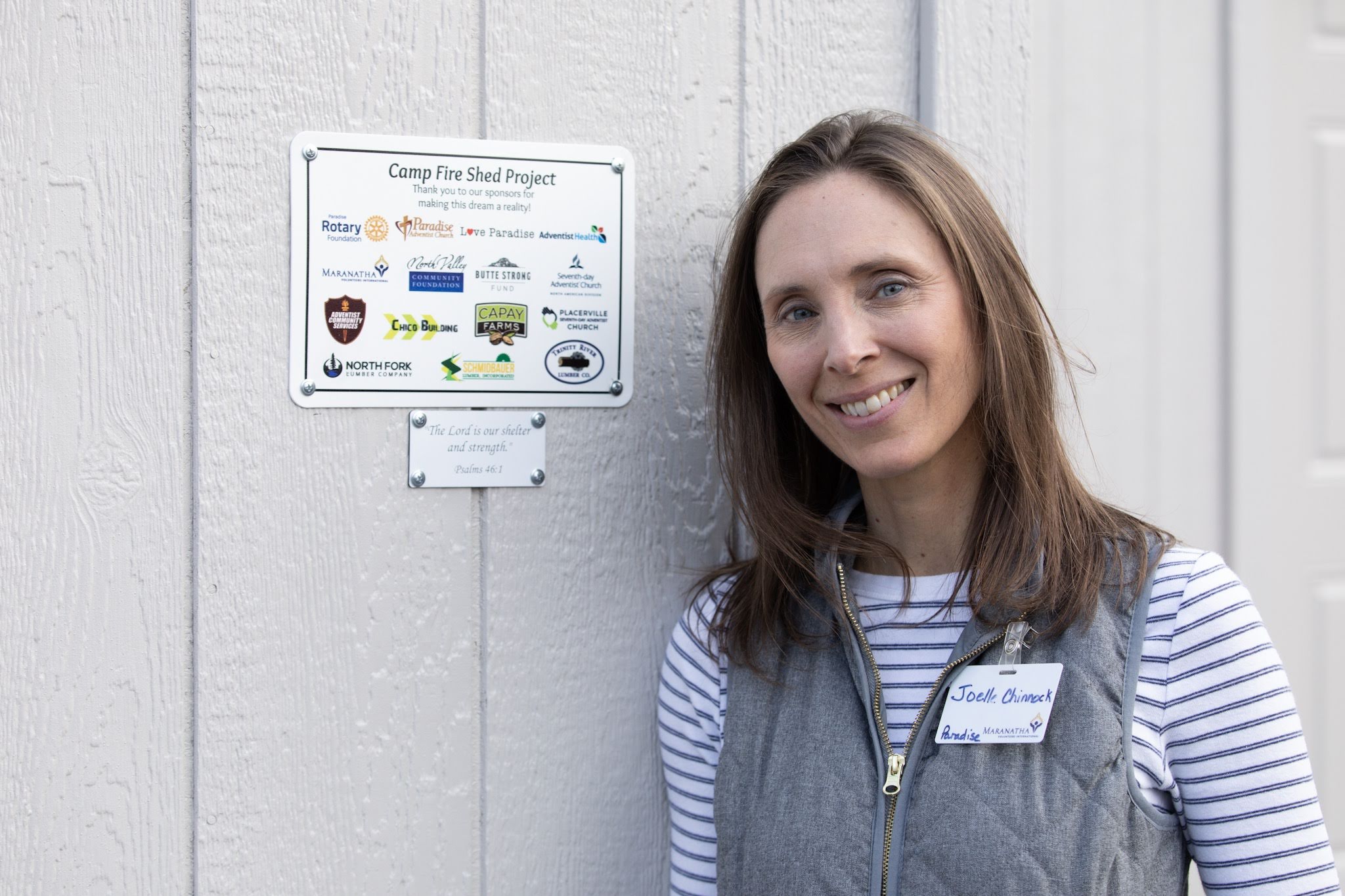 Joelle Chinnock, project coordinator, poses by a finished shed showing the partners that helped make the build possible.