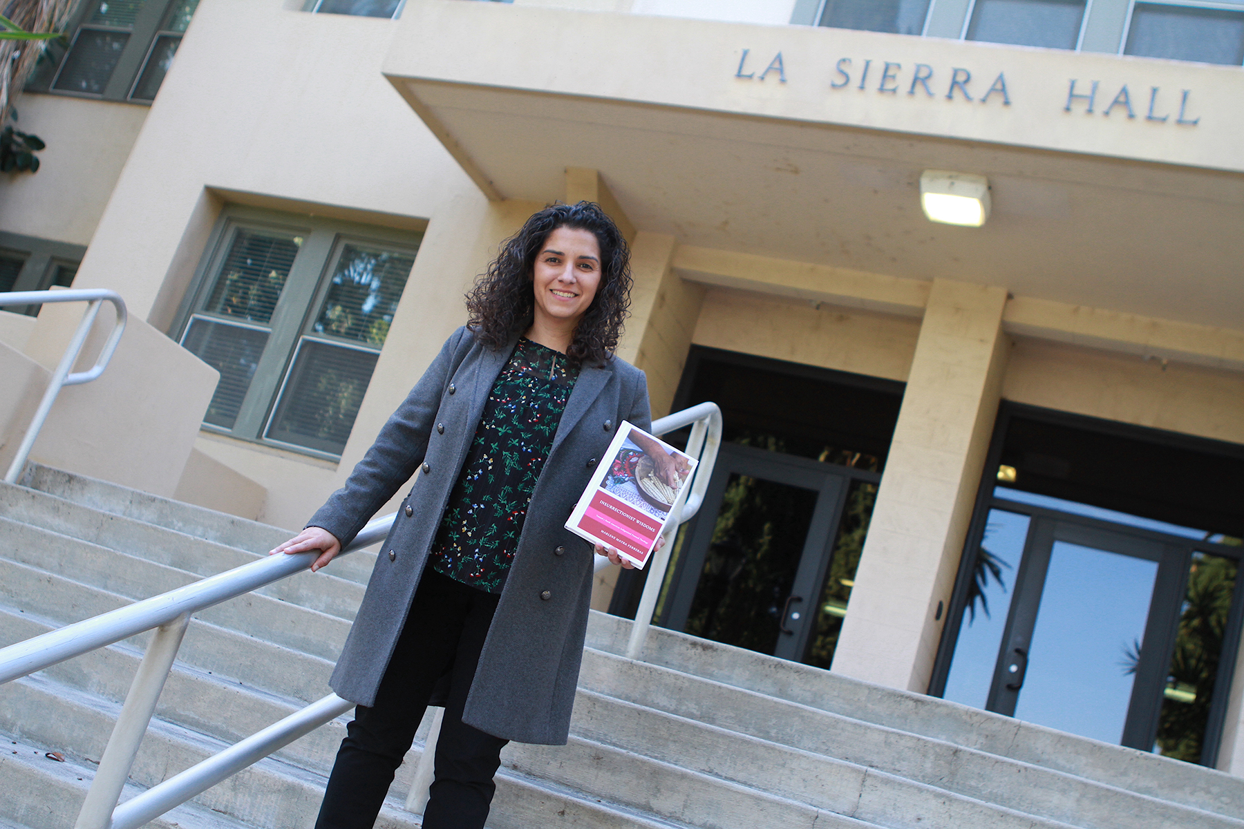 A smiling Hispanic woman stands on the steps of a building named "La Sierra Hall" holding a book. 