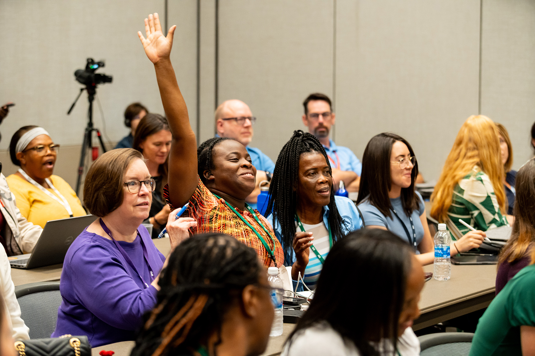 Smiling black woman in a classroom full of people with her hand raised.