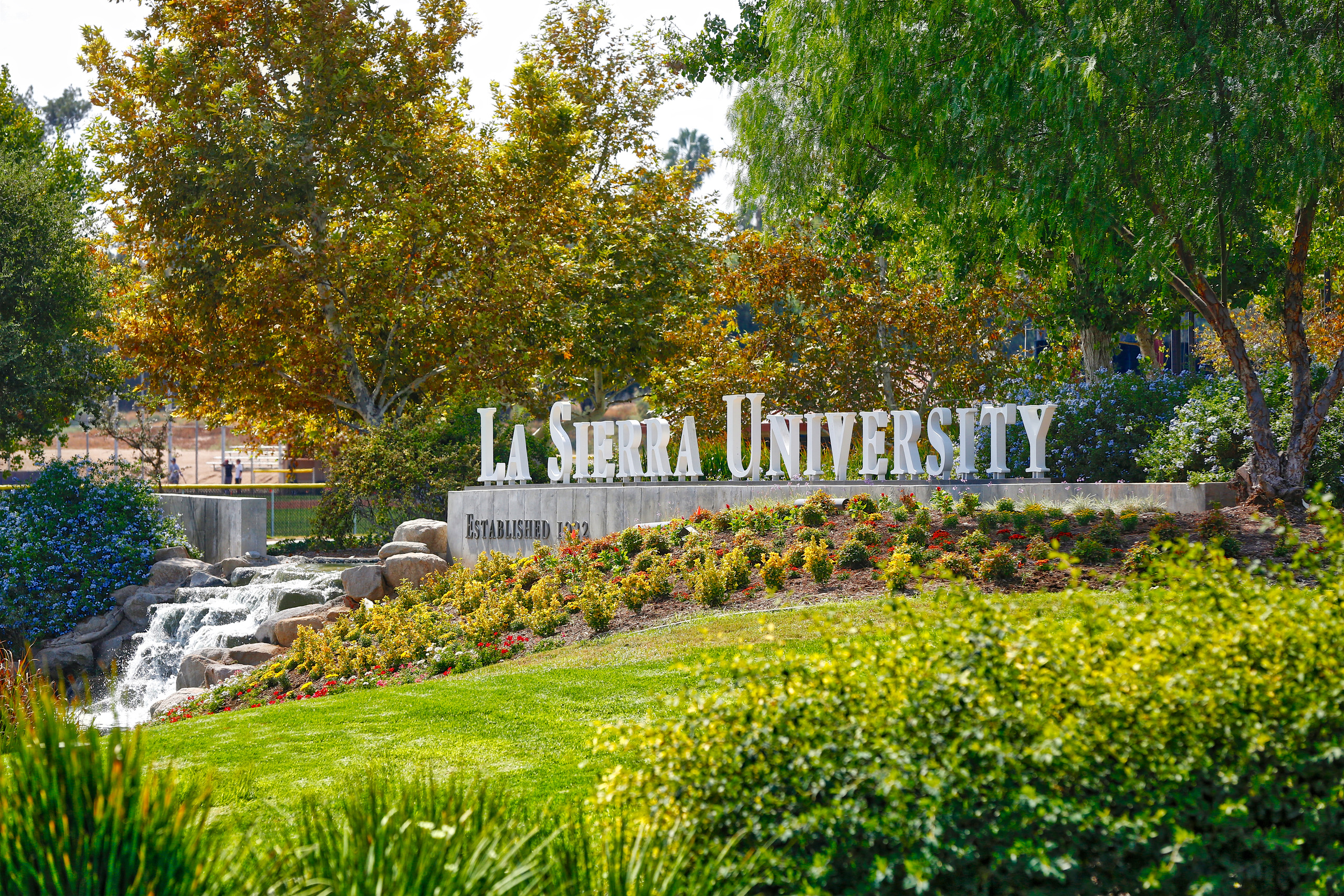 a photo of a sign reading "La Sierra University" surrounded by trees, green grass, plants, and a waterfall. 