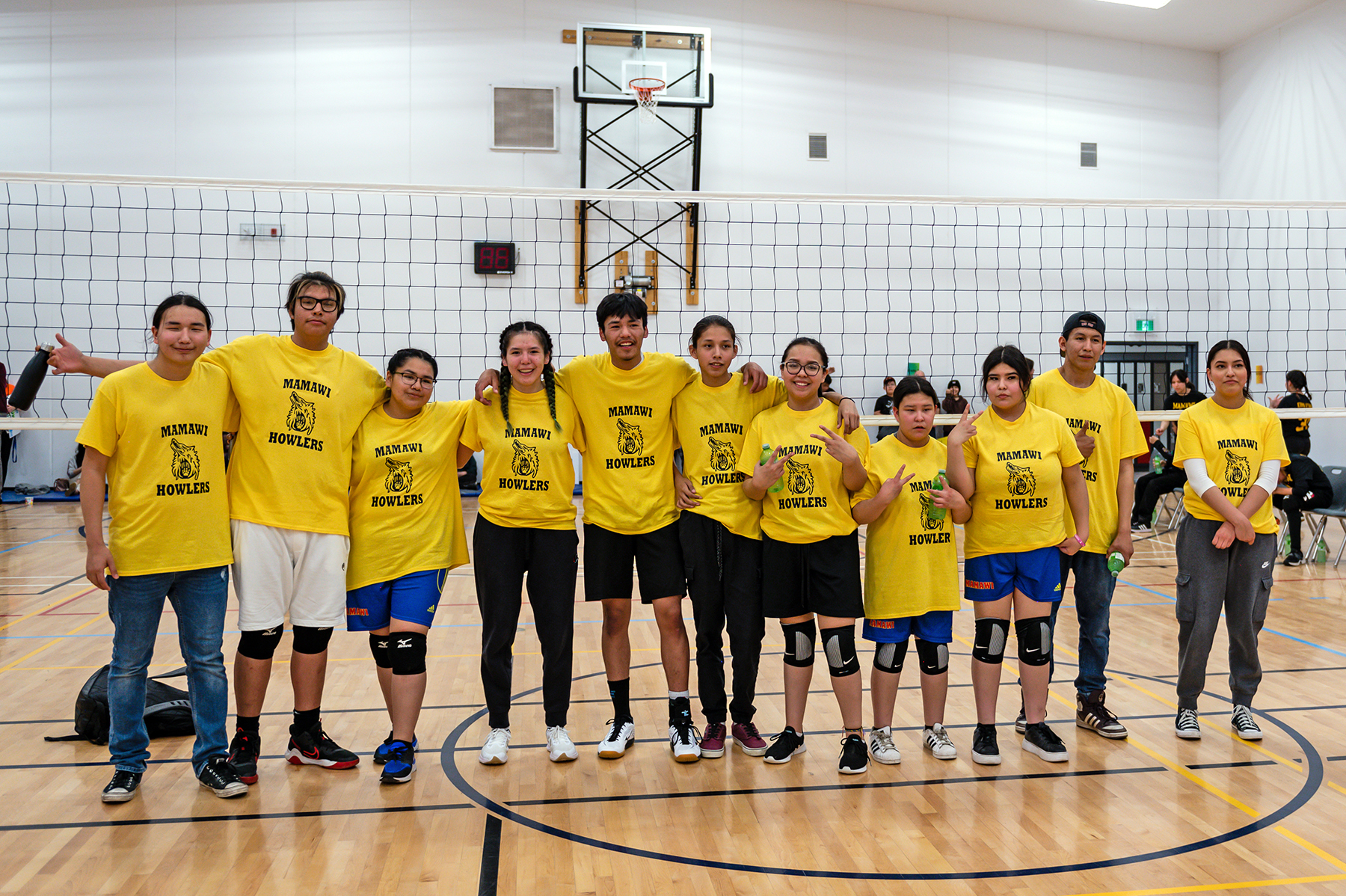 A co-ed group of young people wearing matching yellow shirts reading "Mamawi Howlers" stand in front of a volleyball net