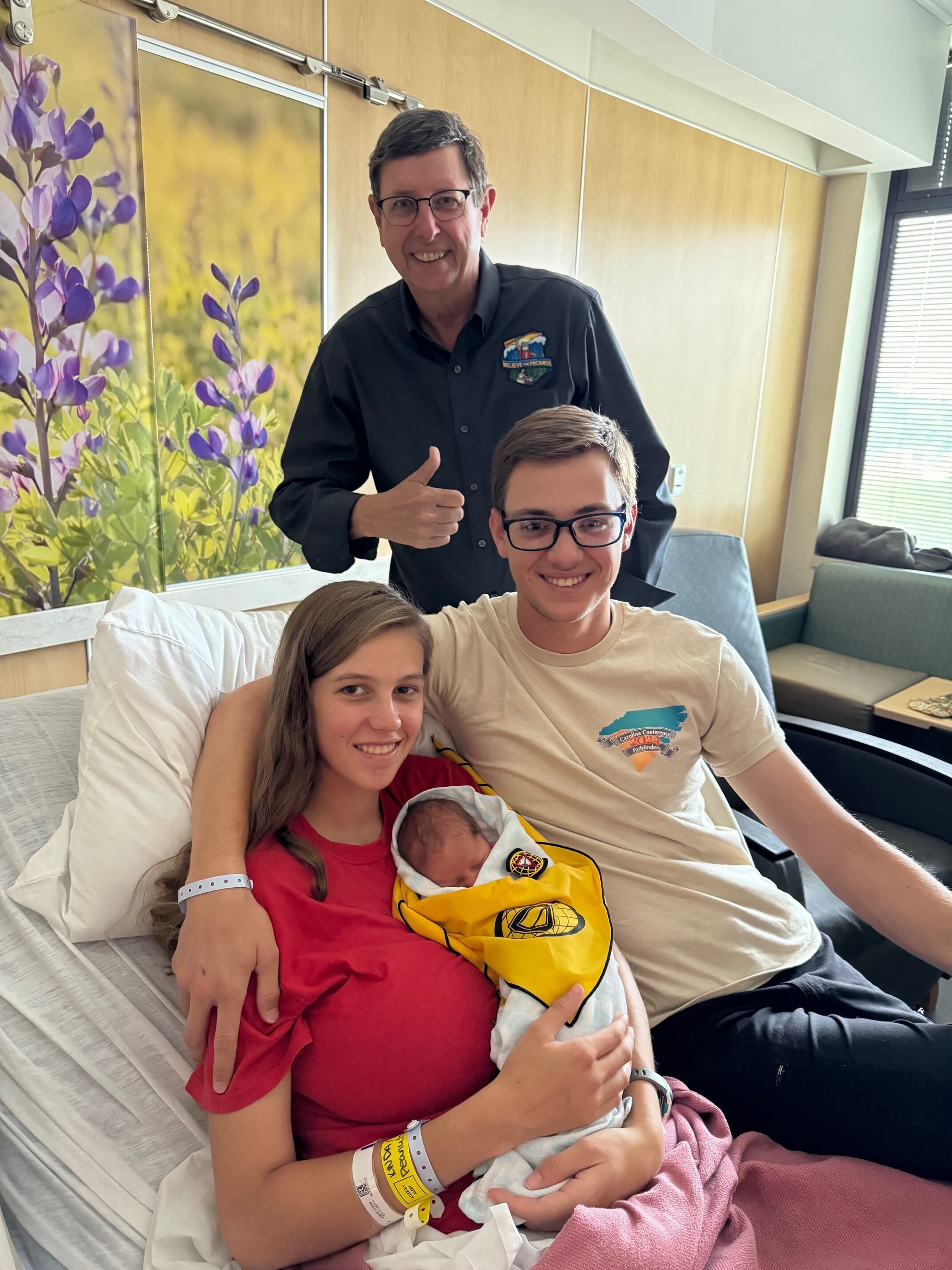 Young adult couple with their newborn baby born during the 2024 Pathfinder camporee in Gillette, Wyoming, posed with middle aged man