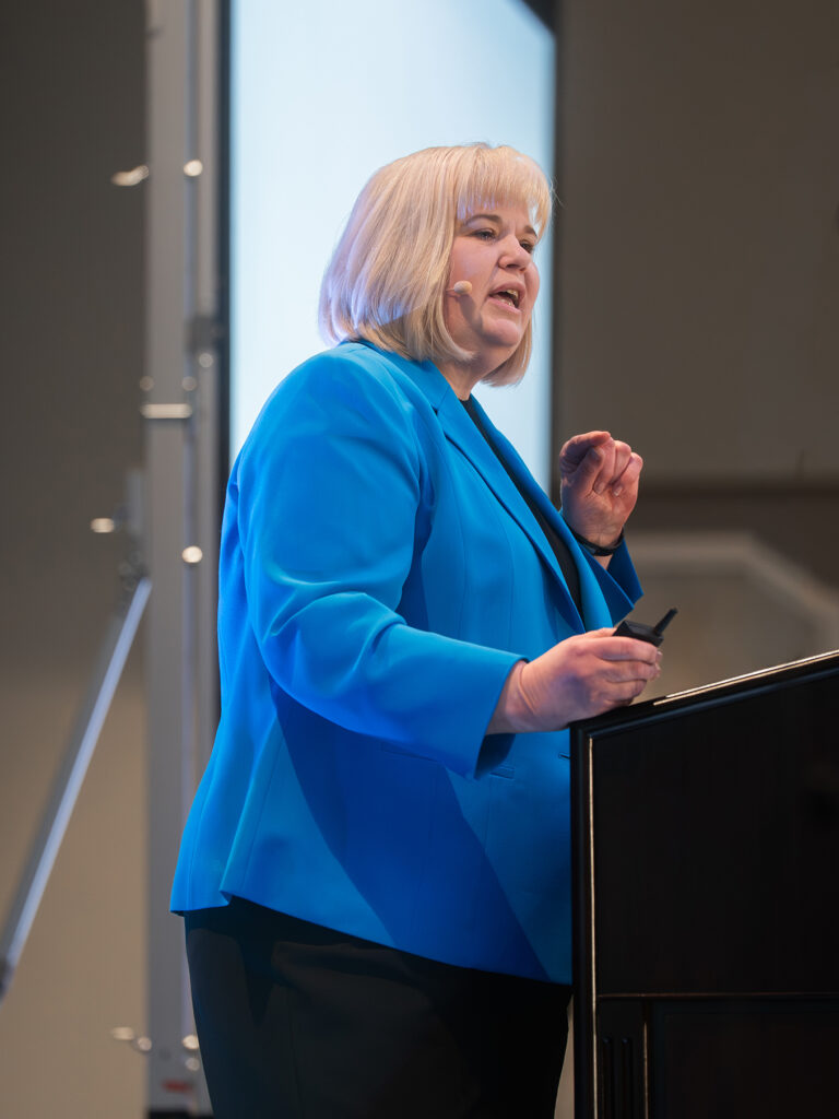 A woman in a blue suit stands at a podium speaking