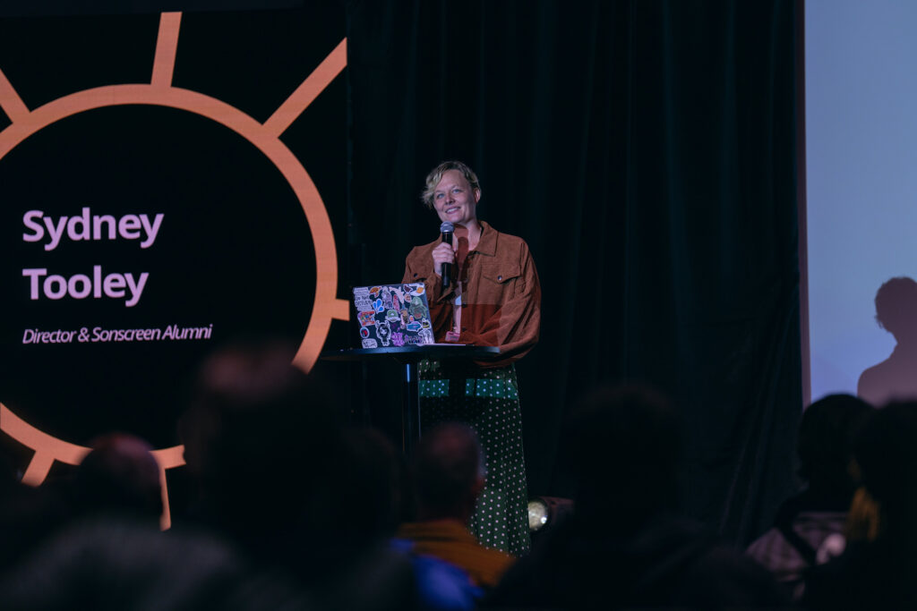 A smiling woman stands on a stage beside a screen describing her as Sydney Tooley, director and Sonscreen alumni. 