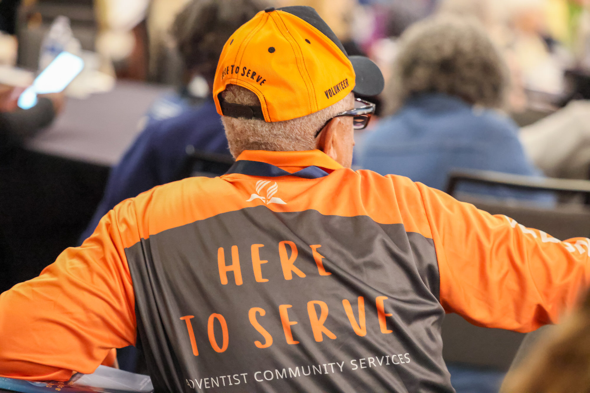 An Adventist Community Services volunteer sitting at a meeting. Photo shows the Here to Serve logo on the back of the shirt and of his hat.