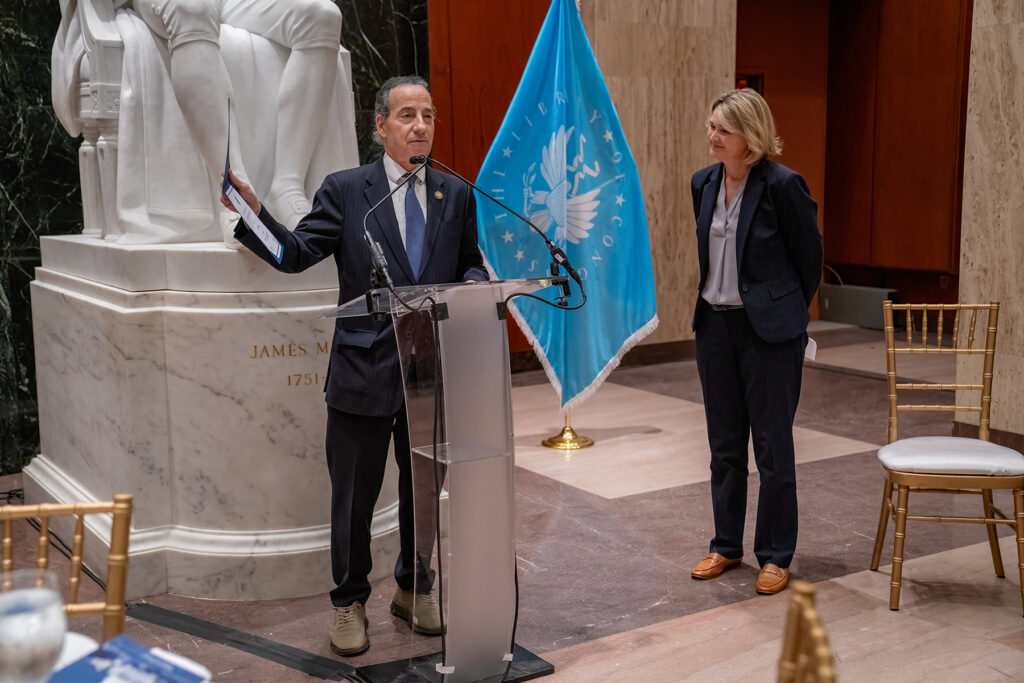 A man speaks at a podium while holding up a certificate, while a woman stands nearby. A Library of Congress flag and statue of James Madison are visible in the background.