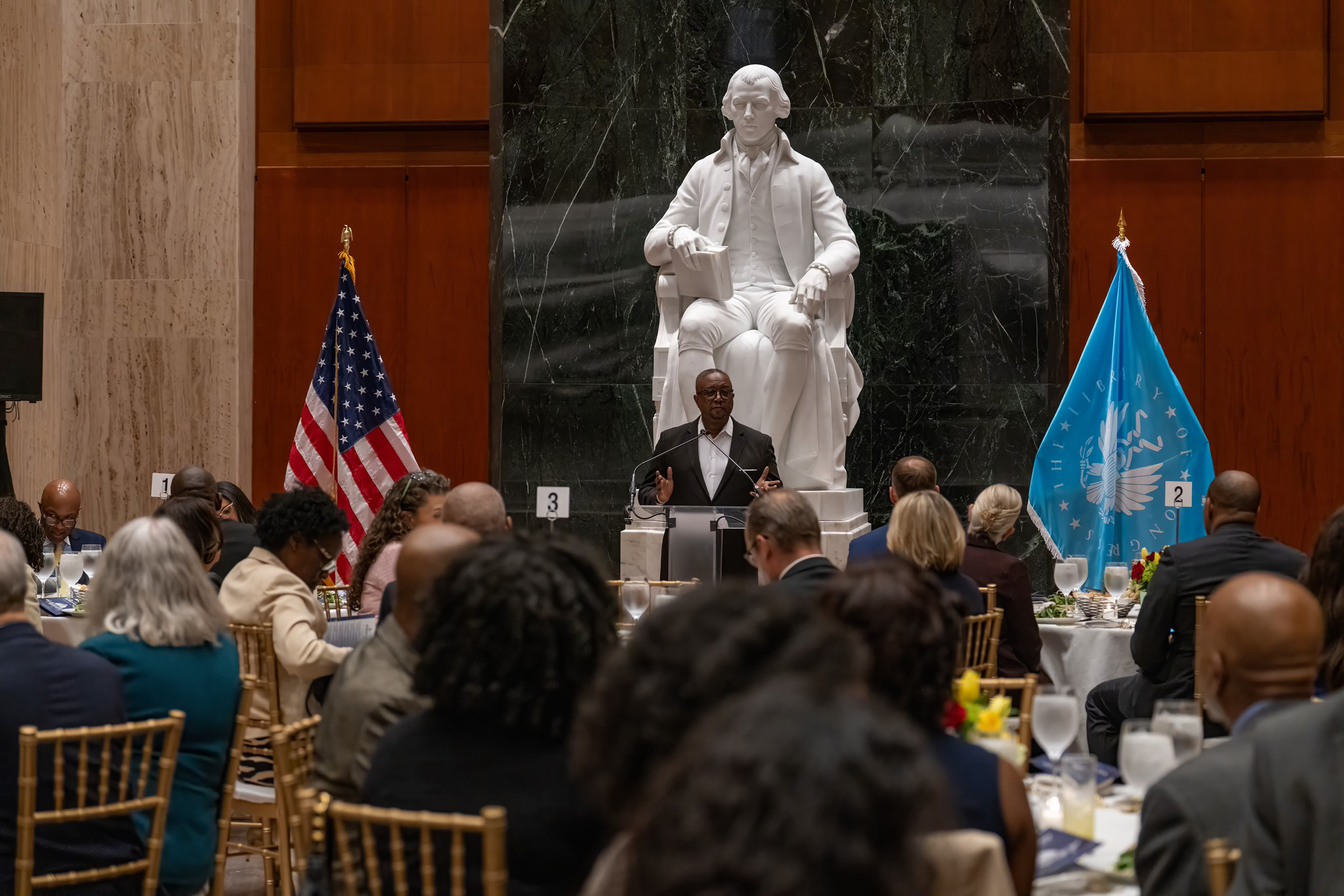 A man stands in front of a crowd speaking with a prominent white statue behind him.