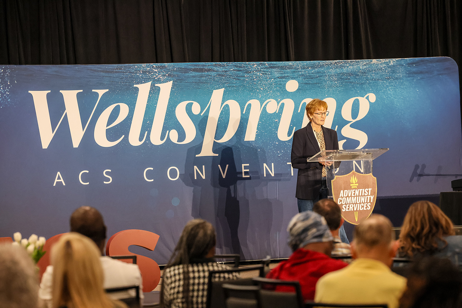 A woman stands in front of an audience at the 2026 "Wellspring" NAD ACS Convention.