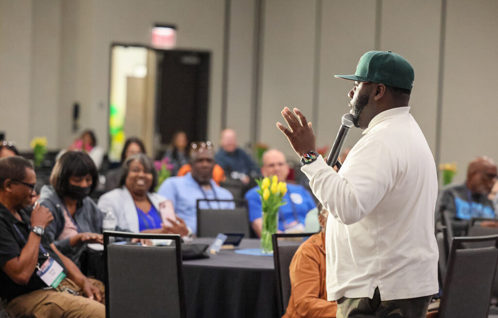 A man in a baseball cap speaks in front of an audience